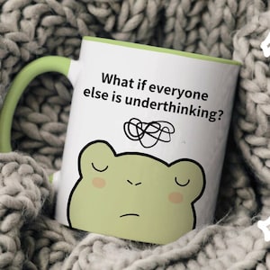 May include: White ceramic mug with a green handle and interior. The mug features a cartoon frog face and the text "What if everyone else is underthinking?" in black. The mug is set against a grey knitted background.