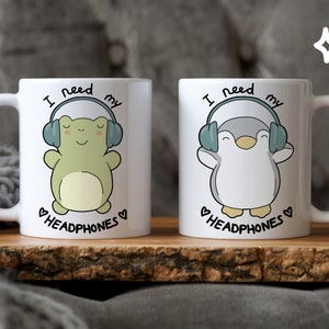 Könnte beinhalten: Zwei weiße Keramikbecher mit Cartoon-Illustrationen. Auf einem Becher ist ein grüner Frosch mit Kopfhörern abgebildet, auf dem anderen ein Pinguin. Beide Becher tragen den Text "I need my headphones" mit einem Herzsymbol.
