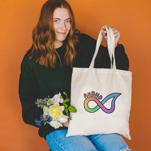 May include: Cream-colored tote bag with the word "AUDHD" in rainbow colors and an infinity symbol design. The bag has long handles and is held up against an orange background.
