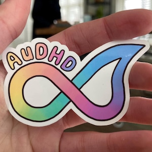Op de afbeelding: Een sticker met het woord "AUDHD" in regenboogkleuren boven een oneindigheidssymbool. Het oneindigheidssymbool is ook regenboogkleurig, met een verloop van geel naar blauw.