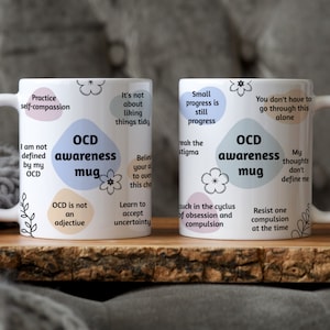 May include: Two white ceramic mugs with the text "OCD awareness mug" and supportive phrases. The mugs are on a wooden tray, with a soft grey background.