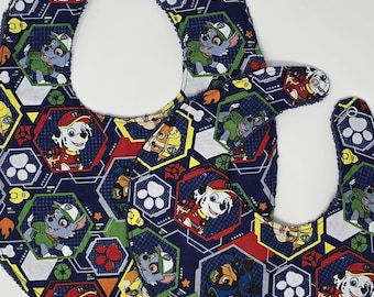 May include: Two blue and white fabric baby bibs with a repeating pattern of cartoon dogs in red, yellow, and green on a blue background. The dogs are wearing different colored hats and have different expressions.