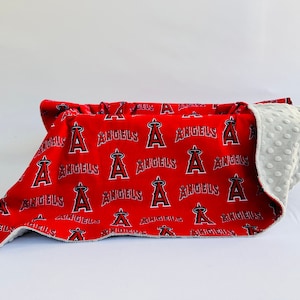 Puede incluir: Una manta para beb&eacute; roja y gris con un patr&oacute;n repetido del logotipo de los &Aacute;ngeles de Los &Aacute;ngeles. La manta est&aacute; doblada por la mitad y el lado gris est&aacute; hacia arriba.