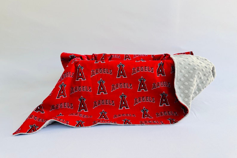 Puede incluir: Una manta para beb&eacute; roja y gris con un patr&oacute;n repetido del logotipo de los &Aacute;ngeles de Los &Aacute;ngeles. La manta est&aacute; doblada por la mitad y el lado gris est&aacute; hacia arriba.