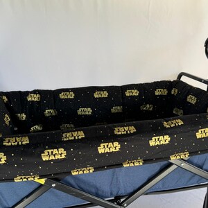 May include: A black and blue wagon with a black frame. The interior lining is black with a gold "Star Wars" logo and star pattern. The wagon has a black handle and wheels.