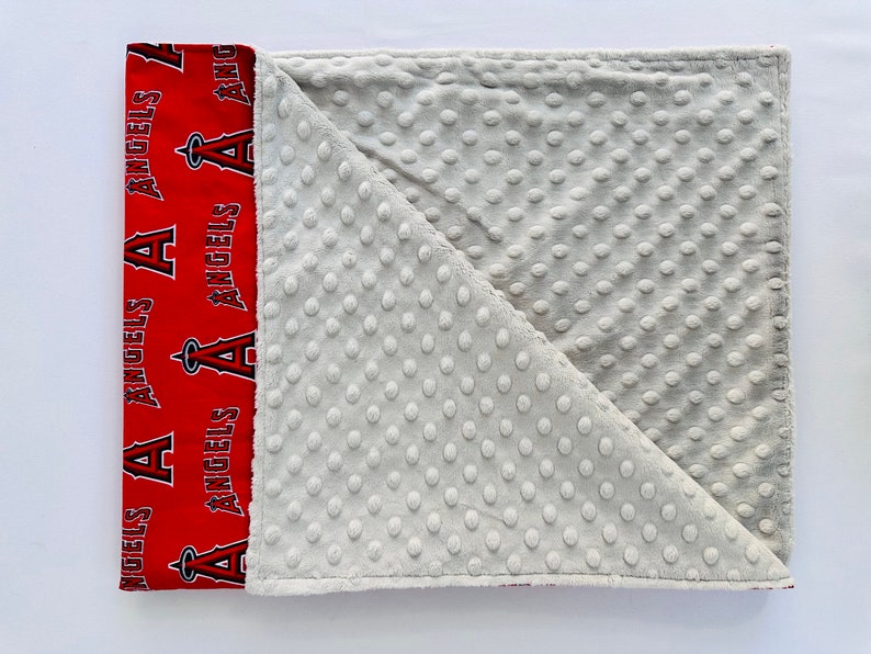 Puede incluir: Una manta para beb&eacute; roja y blanca con un patr&oacute;n de puntos minky gris en un lado y un lado rojo con las palabras "Angels" repetidas.