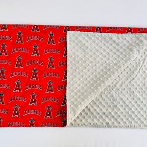 Puede incluir: Una manta para beb&eacute; roja y blanca con un patr&oacute;n del logotipo de los &Aacute;ngeles de Los &Aacute;ngeles. La manta est&aacute; doblada por la mitad y el lado blanco est&aacute; visible. El lado blanco tiene un patr&oacute;n texturizado.
