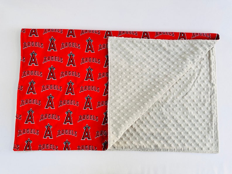 Puede incluir: Una manta para beb&eacute; roja y blanca con un patr&oacute;n del logotipo de los &Aacute;ngeles de Los &Aacute;ngeles. La manta est&aacute; doblada por la mitad y el lado blanco est&aacute; visible. El lado blanco tiene un patr&oacute;n texturizado.