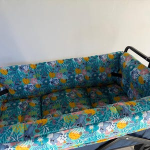 May include: A utility wagon with a padded interior featuring a colorful pattern of koalas, tropical leaves, and yellow fruit on a teal background. The wagon has a black frame and handle.