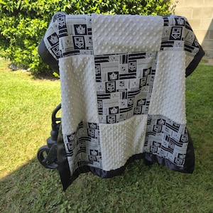 May include: A baby blanket featuring a patchwork design with the Los Angeles Kings logo in black, white, and gray. The blanket has a white, textured, dotted center and a black satin border. It is draped over a stroller.