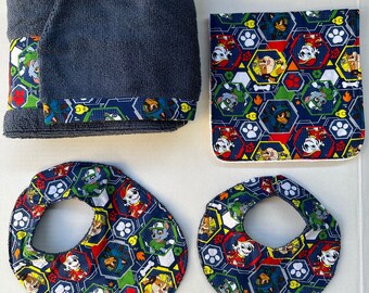 May include: A set of four blue and white fabric items with a paw patrol theme. The set includes a towel, two bibs, and a washcloth. The bibs and washcloth feature a paw patrol character design with a blue background and white paw prints.