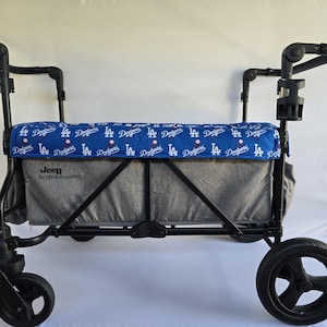 May include: A black Jeep wagon with a gray interior and a blue cover featuring the Los Angeles Dodgers logo. The wagon has black wheels and a cup holder. The text "Jeep by DELTA CHILDREN" is visible on the side.