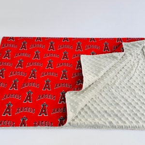 Puede incluir: Una manta para beb&eacute; roja y blanca con un patr&oacute;n repetido del logotipo de los &Aacute;ngeles de Los &Aacute;ngeles. La manta est&aacute; hecha de tela suave y felpa y tiene un respaldo de minky blanco.