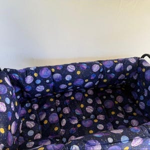 May include: A black metal pet carrier with a padded interior. The interior fabric features a dark blue background with a repeating pattern of purple and yellow planets and stars. The carrier is designed for small pets.