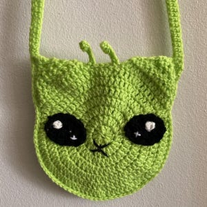 以下が含まれることがあります： ライムグリーンの宇宙人の顔の形をしたかぎ針編みのクロスボディバッグ。バッグは、白いハイライトのある大きな黒い目、小さな触角、ステッチされた口が特徴です。ストラップも同じ緑色の糸でかぎ針編みされています。
