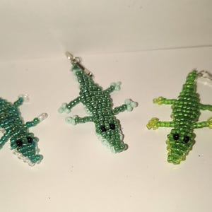 Pode incluir: Três estatuetas de jacaré de contas em tons de verde. Os jacarés têm olhos pretos e são feitos com pequenas contas. Um tem um fecho de chaveiro preso. As estatuetas estão em uma superfície branca.