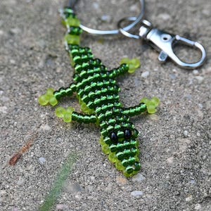 Op de afbeelding: Een kralen sleutelhanger in de vorm van een alligator. De alligator is gemaakt van kleine, donkergroene kralen met lichtgroene poten en accenten. De ogen zijn zwart. De sleutelhanger heeft een zilverkleurige sluiting en ring.