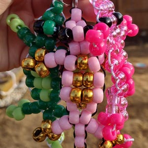 May include: Three beaded keychains in the shape of a dragon. One is pink with gold accents, one is green with gold accents, and one is pink with black accents.