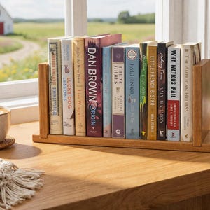 May include: A wooden bookshelf holds a collection of books with colorful spines. The books are arranged on a wooden surface near a window, with a cup of coffee and a succulent plant in the background. The titles are visible.