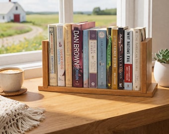 May include: A wooden bookshelf holds a collection of books with colorful spines. The books are arranged on a wooden surface near a window, with a cup of coffee and a succulent plant in the background. The titles are visible.