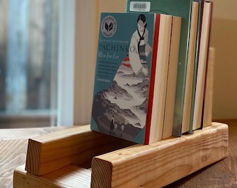 May include: A wooden book stand holds several books, including one titled "Pachinko" by Min Jin Lee. The stand is made of light-colored wood and has a simple, rectangular design. The books are stacked on a wooden table.