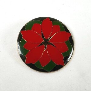 May include: A circular enamel pin featuring a vibrant red poinsettia flower with dark green leaves set against a black background. The design is outlined in thin gold-toned metal, creating a defined contrast between the floral petals and the dark circular border of the accessory.