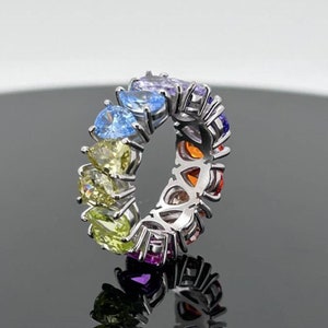 May include: A silver ring with a rainbow of colorful gemstones set in a prong setting. The ring is made of metal and has a polished finish.