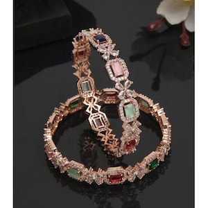 May include: A pair of rose gold bangles with intricate designs featuring clear and colored gemstones. The bangles are adorned with floral motifs and rectangular-shaped stones in shades of green, pink, red, and blue.