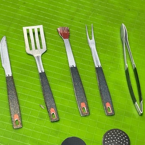 May include: A set of miniature barbecue tools, including a knife, spatula, brush, fork, and tongs. The tools have silver blades and black handles with red accents. The items are arranged on a bright green surface.