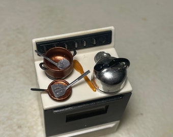 May include: A miniature white stovetop with a black oven door. There are two copper pots with handles and a silver kettle with a black handle on the stovetop. A wooden spoon and a metal spatula are also on the stovetop.