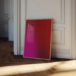 May include: A framed art print with a gradient of red to magenta hues. The artwork leans against a white wall and a wooden floor. The frame is a light wood color. The artwork reflects the light from a window.