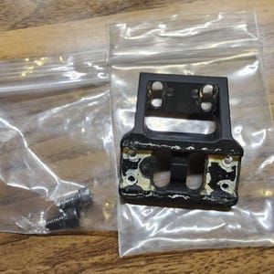 Op de afbeelding: Een zwarte metalen montageplaat met vier schroefgaten en een rechthoekige uitsparing. De plaat zit in een doorzichtige plastic zak met twee kleine schroeven.