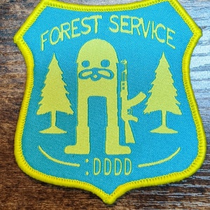 May include: A yellow and green embroidered patch featuring a cartoon figure holding a rifle, two pine trees, and the text "FOREST SERVICE : DDDD".