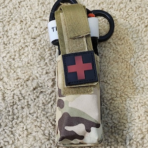 May include: A camouflage-patterned medical pouch with a red cross emblem. The pouch contains a tourniquet and is secured with a black ring and tan straps. The text "TI" is visible on a white strap.