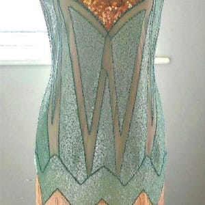 May include: A vintage-style flapper dress on a mannequin. The dress features a V-neck, intricate sequin detailing in gold and teal, and a fringed hemline. The dress is a classic representation of the roaring twenties.