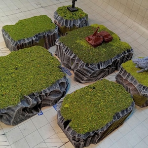 Puede incluir: Cinco bases de terreno rocoso grises y negras con hierba verde en la parte superior. Las bases son de diferentes tamaños y formas. Una base tiene un pequeño vehículo gris en la parte superior, otra tiene una criatura gris en la parte superior.