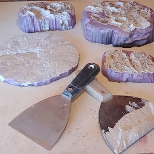May include: An assortment of purple and white textured foam pieces, possibly for crafting or model building, are arranged on a light-colored surface. Two metal putty knives with black and wooden handles are also present.