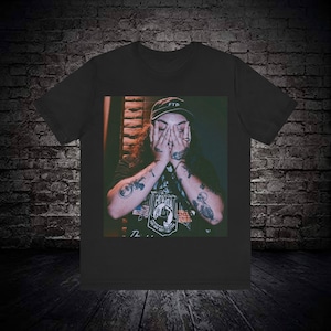 May include: Black t-shirt with a graphic image of a person with tattoos, wearing a hat with "FTP" on it. The person is covering their face with their hands. The shirt has a vintage, slightly faded appearance.