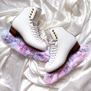 May include: A pair of white figure skates with gold accents and a brown wooden base. The skates are on top of a pair of purple and blue fuzzy skate guards.