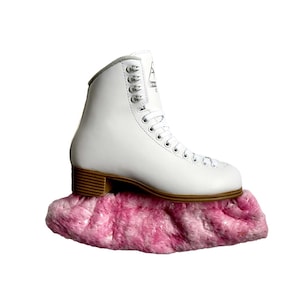 May include: A white figure skate with silver eyelets and a brown heel sits atop a fluffy pink skate guard. The skate has a high-top design and a logo on the side. The skate guard is a soft, plush material.