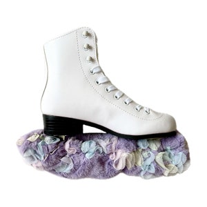 May include: A white ice skate with black heels and white laces rests on a lavender faux fur skate guard. The skate guard is decorated with pastel-colored fabric flowers in shades of blue, pink, and white. The image is set against a white background.