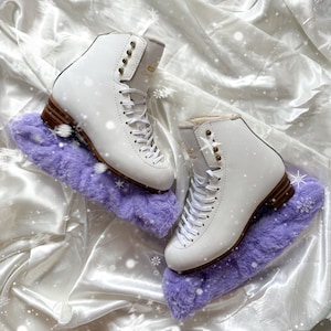 May include: Two white figure skates with purple fuzzy boot covers. The skates have gold accents and laces. The skates are on a white satin background.