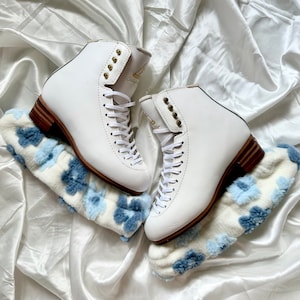 Op de afbeelding: Een paar witte schaatsen met bruine hakken en goudkleurige oogjes. De schaatsen zijn geregen en liggen op een blauw en wit bloemmotief accessoire. Op een witte satijnen achtergrond.