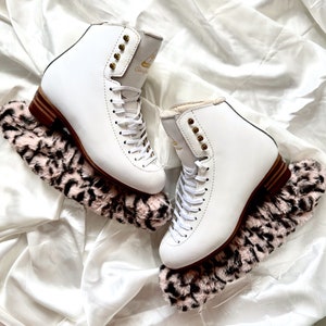 May include: A pair of white figure skates with gold accents and black laces. The skates are on top of pink and black faux fur boot covers.