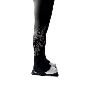 以下が含まれることがあります： 脚の下部にスタイリッシュな鳥のラインストーンのデザインが施された黒いフィギュアスケートパンツ。パンツは白いフィギュアスケート靴と合わせています。