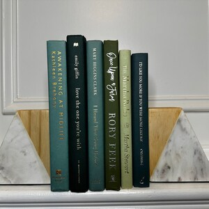 May include: A set of bookends made of white marble and wood, holding up a row of seven books with green and blue covers. The book titles are visible, including "Awakening at Midlife", "Love the One You're With", "I Heard That Song Before", "Once Upon a Farm", "The Martha Rules", and "I'd Like You More If You Were More Like Me".