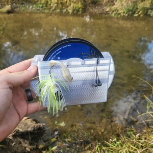 May include: A clear plastic fishing lure storage box with two spools of fishing line and a lure with a white and green feather tail.