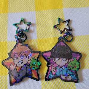 May include: Two iridescent star-shaped keychains featuring anime characters. The keychain on the left depicts a character with brown hair and a green ghost. The keychain on the right shows a character with black hair and a green ghost. Both keychains have a rainbow iridescent finish.