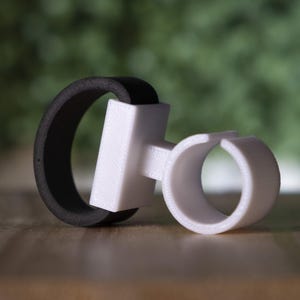 May include: A white and black 3D printed phone ring holder. The ring holder has a black rubber band and a white plastic base with a circular opening.