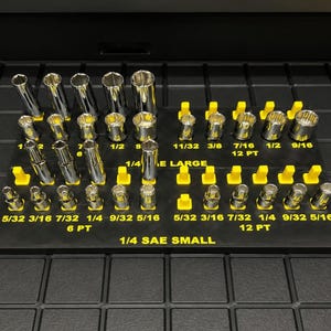 May include: A black socket organizer with yellow holders, filled with various sizes of silver sockets. The text "1/4 SAE SMALL" and "1/4 SAE LARGE" is visible. The sockets are arranged in rows, with sizes labeled in inches.